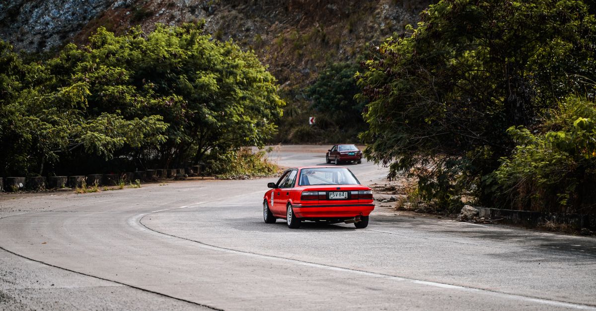 A red car driving on a scenic mountain road in Mangatarem, Philippines, surrounded by greenery and hills.